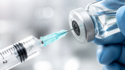 Close-up of a syringe needle piercing the rubber stopper of a clear pharmaceutical vial held with a gloved hand, illustrating medication preparation, injectable drug administration, or sterile laboratory testing.