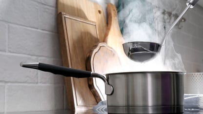 Adding water with ladle into steaming saucepan on stove in kitchen